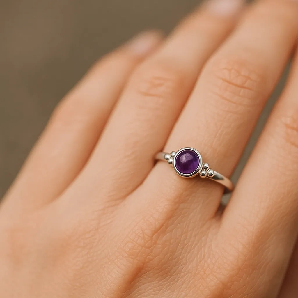Eine Nahaufnahme einer Frauenhand zeigt einen eleganten Silberring mit einem runden, violett schimmernden Amethyst-Cabochon. Der glatte, 4 mm große Stein ist in eine schlichte Fassung eingefasst und wird von kleinen silbernen Kugeldetails flankiert. Natürliches, weiches Licht hebt den Glanz des Silbers und die Tiefe des violetten Steins hervor, während der neutrale Hintergrund den Fokus auf den Ring lenkt.