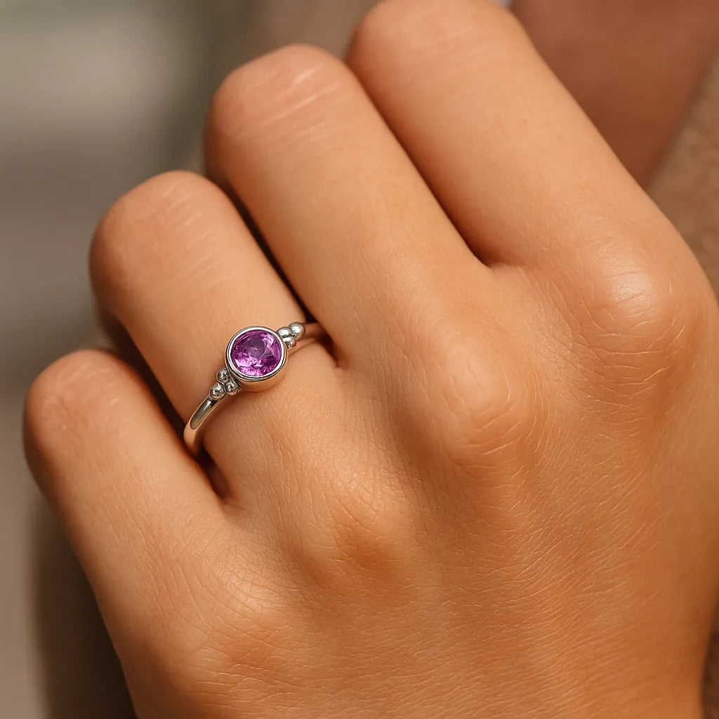 Eine Nahaufnahme einer Frauenhand zeigt einen eleganten Silberring mit einem runden, facettierten Amethyst. Der violette Edelstein mit 4 mm Durchmesser ist in eine glatte Silberfassung eingefasst und wird von kleinen Kugeldetails flankiert. Natürliches, weiches Licht hebt den Glanz des Steins und der polierten Ringschiene hervor, während der neutrale Hintergrund den Fokus auf das Schmuckstück lenkt.
