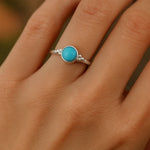 Eine Nahaufnahme einer Frauenhand zeigt einen eleganten Silberring mit einem runden, himmelblauen Chalcedon-Cabochon. Der 4 mm große Stein ist glatt poliert und in eine schlichte Silberfassung mit kleinen Kugeldetails eingefasst. Natürliches Licht hebt den sanften Glanz des Steins und der Ringschiene hervor, während der neutrale Hintergrund den Fokus auf den Ring lenkt.
