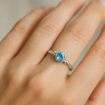 Eine Nahaufnahme einer Frauenhand zeigt einen eleganten Silberring mit einem runden, facettierten Blautopas. Der hellblaue Edelstein mit 4 mm Durchmesser ist in eine glatte Fassung eingefasst und wird von kleinen silbernen Kugeldetails flankiert. Natürliches, weiches Licht betont den klaren Glanz des Steins und die polierte Oberfläche der Ringschiene vor einem neutralen, unscharfen Hintergrund.
