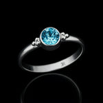 Silberring mit rundem blauem Topas in Zargenfassung, liegend auf schwarzem spiegelndem Untergrund mit klarer Reflexion, edles Schmuck Produktfoto in leicht schräger Perspektive.