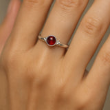 Nahaufnahme einer Frauenhand mit einem eleganten Silberring, der einen tiefroten, runden Granat-Cabochon als Mittelstein trägt. Der 4 mm große Edelstein ist in eine glatte Fassung eingebettet und reflektiert sanft das Licht. Der Fokus des Bildes liegt auf dem Ring, während der Hintergrund neutral und weich gehalten ist, um den Glanz des Silbers und die warme Farbe des Granats hervorzuheben.
