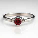 `Hochwertiges Produktfoto eines filigranen Damenrings aus 925 Sterling Silber mit rund facettiertem Granat (4 mm) in Zargenfassung und seitlichen Silberkügelchen, frontal aufgenommen auf hellem spiegelnden Hintergrund 