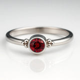 `Hochwertiges Produktfoto eines filigranen Damenrings aus 925 Sterling Silber mit rund facettiertem Granat (4 mm) in Zargenfassung und seitlichen Silberkügelchen, frontal aufgenommen auf hellem spiegelnden Hintergrund 