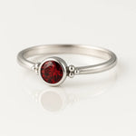 `Filigraner Damenring aus 925 Sterling Silber mit rund facettiertem Granat (4 mm) in Zargenfassung und kleinen Silberkügelchen an den Seiten, edel fotografiert auf hellem spiegelnden Untergrund als hochwertiges Schmuck-Produktfoto.`
