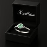 Eleganter Silberring mit rund facettiertem grünem Edelstein (4 mm) in Zargenfassung, präsentiert in geöffneter schwarzer Schmuckbox mit Novellara Logo – hochwertiger Damenring als Geschenk oder Verlobungsring.
