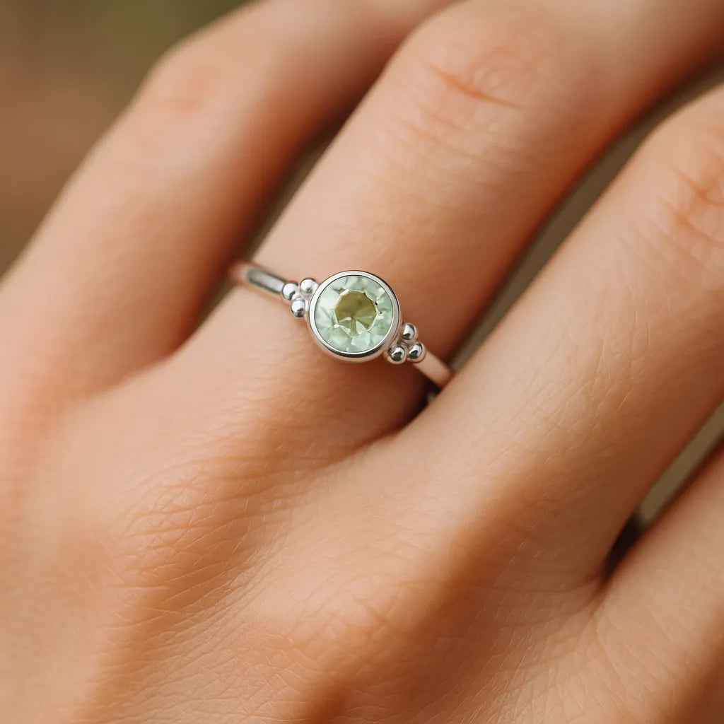 Eine Nahaufnahme einer Frauenhand zeigt einen filigranen Silberring mit einem runden, facettierten Grünquarz. Der zartgrüne, leicht transparente Stein mit 4 mm Durchmesser ist in eine glatte Silberfassung eingefasst und wird von kleinen silbernen Kugeldetails flankiert. Weiches, natürliches Licht betont den feinen Glanz des Rings und die dezente Farbe des Edelsteins vor einem neutralen Hintergrund.

