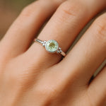 Eine Nahaufnahme einer Frauenhand zeigt einen filigranen Silberring mit einem runden, facettierten Grünquarz. Der zartgrüne, leicht transparente Stein mit 4 mm Durchmesser ist in eine glatte Silberfassung eingefasst und wird von kleinen silbernen Kugeldetails flankiert. Weiches, natürliches Licht betont den feinen Glanz des Rings und die dezente Farbe des Edelsteins vor einem neutralen Hintergrund.
