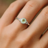 Eine Nahaufnahme einer Frauenhand zeigt einen filigranen Silberring mit einem runden, facettierten Grünquarz. Der zartgrüne, leicht transparente Stein mit 4 mm Durchmesser ist in eine glatte Silberfassung eingefasst und wird von kleinen silbernen Kugeldetails flankiert. Weiches, natürliches Licht betont den feinen Glanz des Rings und die dezente Farbe des Edelsteins vor einem neutralen Hintergrund.
