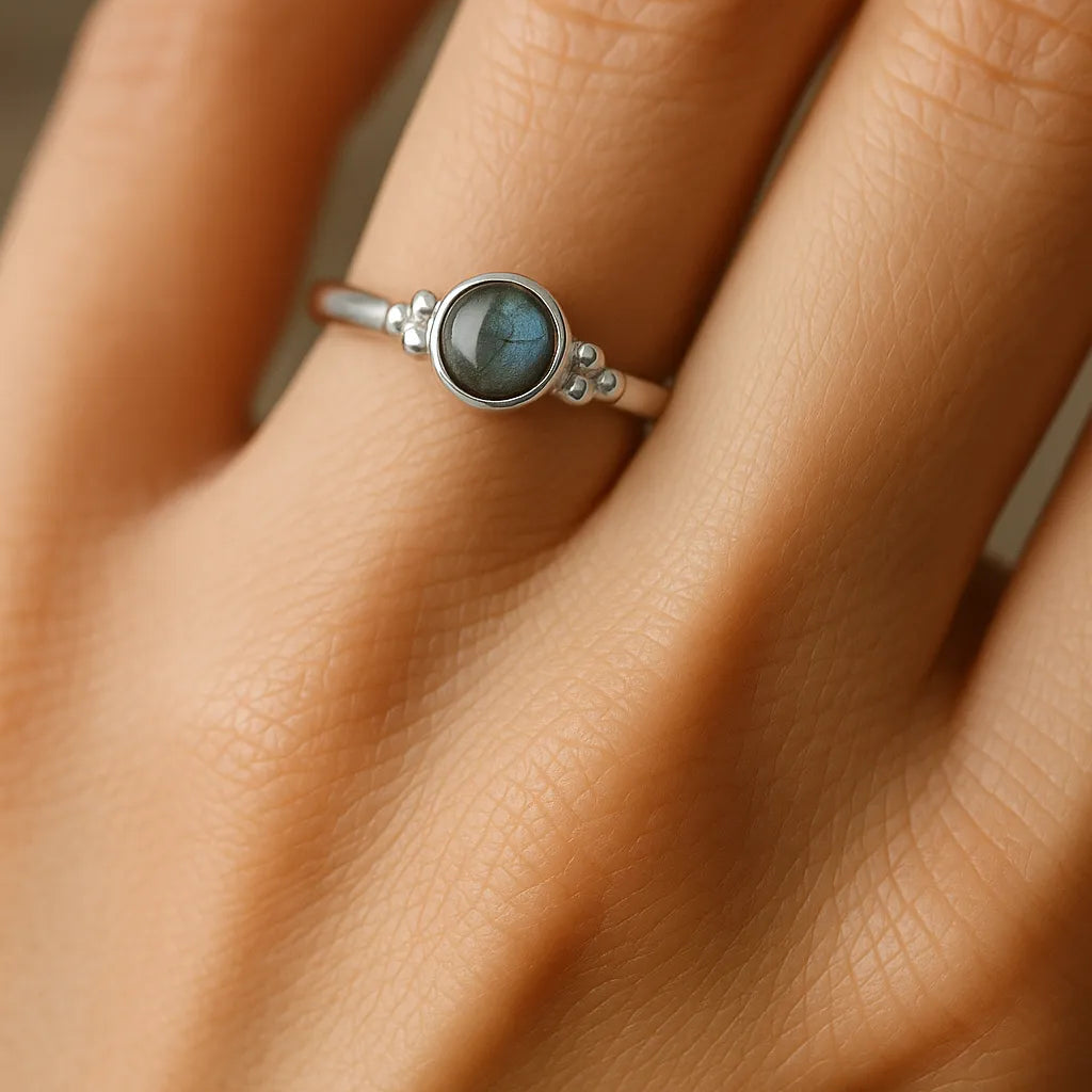 „Nahaufnahme einer Frauenhand, die einen eleganten Silberring mit einem runden Labradorit-Cabochon trägt. Der graublaue Stein zeigt ein sanftes Schimmern in Grün- und Blautönen und ist in eine feine, glänzende Ringschiene eingefasst. Der Fokus liegt auf dem Ring, der in natürlichem Licht dezent reflektiert.“