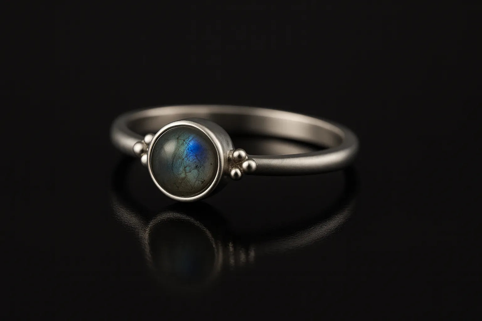 Damenring aus Sterling Silber mit rundem Labradorit Cabochon (4 mm) in Zargenfassung – filigraner Silberring mit blau-grün schimmerndem Edelstein, edel inszeniert auf schwarzem spiegelnden Hintergrund in seitlicher Perspektive.
