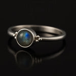 Damenring aus Sterling Silber mit rundem Labradorit Cabochon (4 mm) in Zargenfassung – filigraner Silberring mit blau-grün schimmerndem Edelstein, edel inszeniert auf schwarzem spiegelnden Hintergrund in seitlicher Perspektive.
