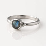 Damenring aus Sterling Silber mit rundem Labradorit Cabochon (4 mm) in Zargenfassung – eleganter Silberring mit blau-grün schimmerndem Edelstein, fotografiert seitlich auf weißem spiegelnden Hintergrund als exklusiver Damenschmuck.
