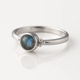 Damenring aus Sterling Silber mit rundem Labradorit Cabochon (4 mm) in Zargenfassung – eleganter Silberring mit blau-grün schimmerndem Edelstein, fotografiert seitlich auf weißem spiegelnden Hintergrund als exklusiver Damenschmuck.

