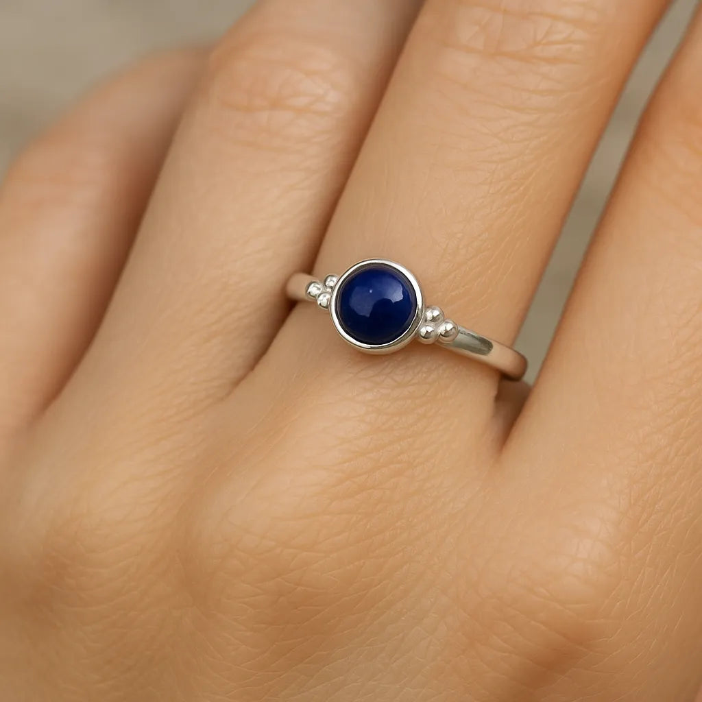 Eine Nahaufnahme einer Frauenhand, die einen eleganten Silberring mit einem kleinen, runden Lapislazuli-Cabochon trägt. Der tiefblaue Stein ist glatt poliert und in eine schlichte Silberfassung mit feinen Kugeldetails eingefasst. Der Fokus liegt auf dem Ring, während der Hintergrund neutral und weich beleuchtet ist.
