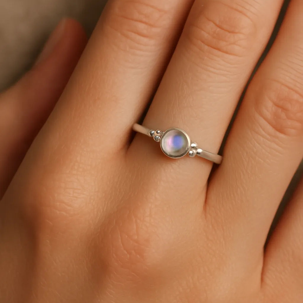 Nahaufnahme einer Frauenhand mit einem eleganten Silberring am Ringfinger. Der Ring zeigt einen etwa 4 mm großen, runden, facettierten Mondstein in einer glatten Zargenfassung, flankiert von kleinen Silberkügelchen. Der Mondstein schimmert mit einem zarten bläulichen Glanz, während das weiche Licht die natürliche Hautstruktur und den sanften Glanz des Silbers betont.
