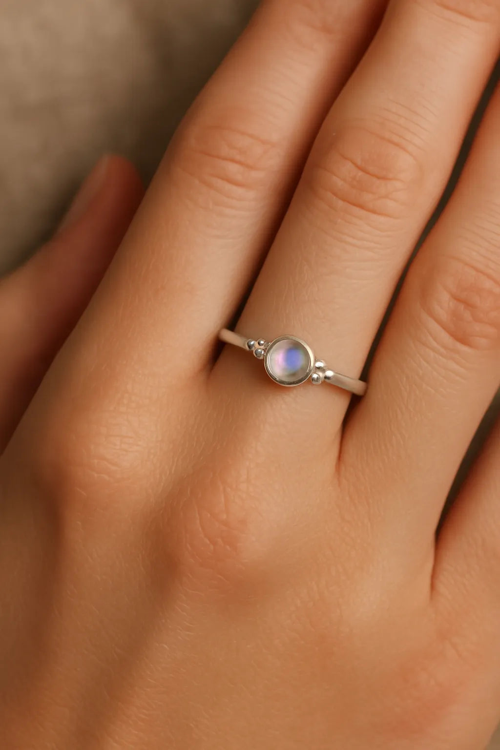 Nahaufnahme einer Frauenhand mit einem eleganten Silberring am Ringfinger. Der Ring zeigt einen etwa 4 mm großen, runden, facettierten Mondstein in einer glatten Zargenfassung, flankiert von kleinen Silberkügelchen. Der Mondstein schimmert mit einem zarten bläulichen Glanz, während das weiche Licht die natürliche Hautstruktur und den sanften Glanz des Silbers betont.
