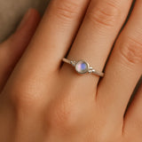 Nahaufnahme einer Frauenhand mit einem eleganten Silberring am Ringfinger. Der Ring zeigt einen etwa 4 mm großen, runden, facettierten Mondstein in einer glatten Zargenfassung, flankiert von kleinen Silberkügelchen. Der Mondstein schimmert mit einem zarten bläulichen Glanz, während das weiche Licht die natürliche Hautstruktur und den sanften Glanz des Silbers betont.
