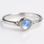 Zarter Silberring mit rundem Regenbogenmondstein in Zargenfassung, liegend auf hellem reflektierendem Untergrund, Produktfoto mit leichter seitlicher Perspektive für modernen Schmuck