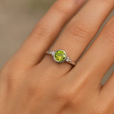 Eine Nahaufnahme einer Frauenhand zeigt einen silbernen Ring mit einem glatten, hellgrünen Peridot-Stein. Der Fokus liegt auf dem Ring, der im weichen, natürlichen Licht schimmert. Der Hintergrund ist neutral und unscharf, wodurch der edle Glanz des Silbers und die klare Farbe des Steins besonders betont werden.