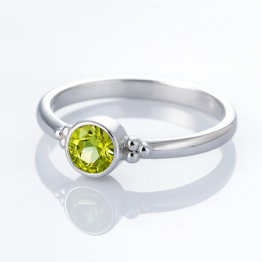 Schlichter Silberring mit einem runden, facettierten, hellgrün leuchtenden Stein (Peridot) in einer runden Fassung, seitlich mit kleinen silbernen Kügelchen verziert, auf weißem Hintergrund liegend und spiegelnd.
