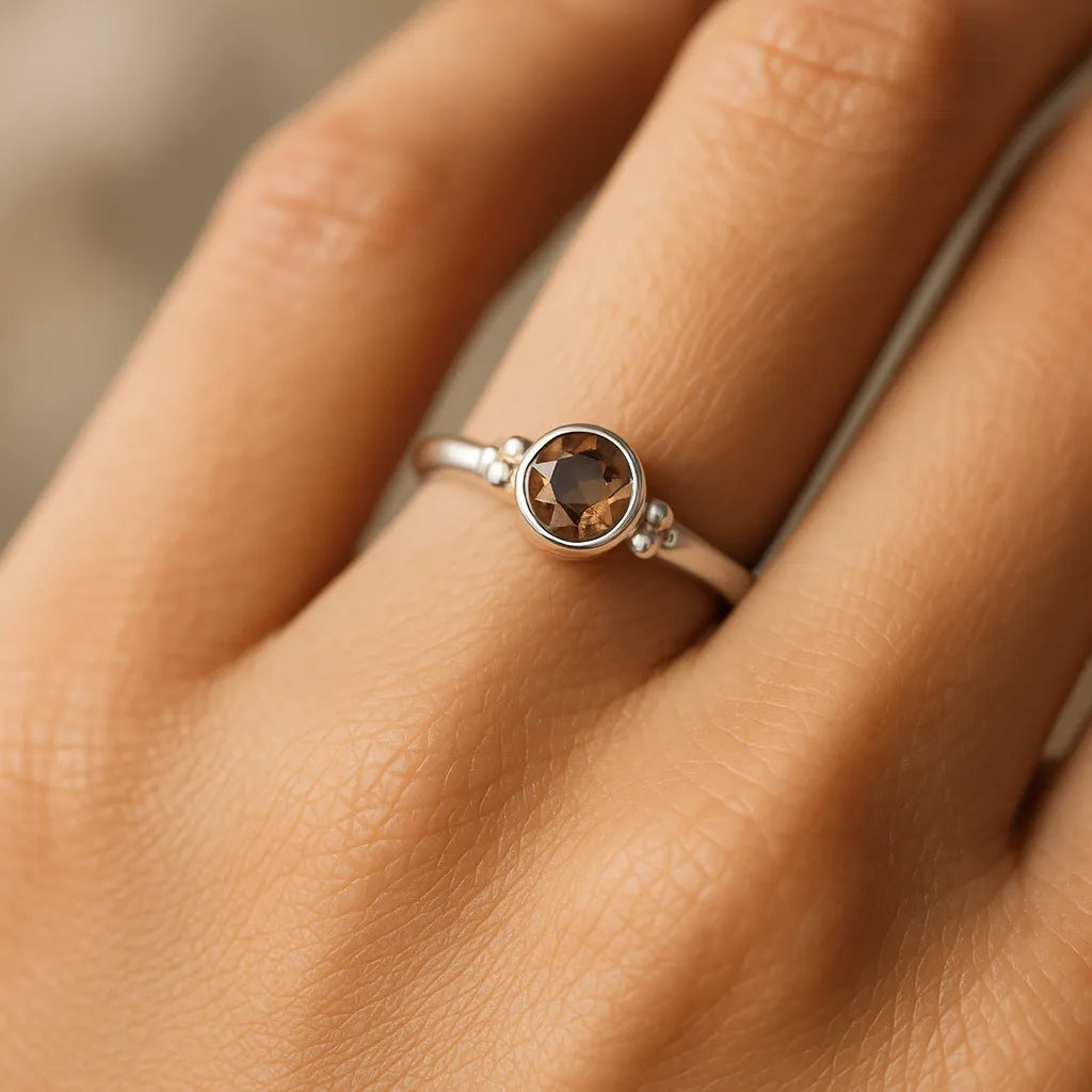Eine Nahaufnahme einer Frauenhand zeigt einen eleganten Silberring mit einem runden, facettierten Rauchquarz. Der 4 mm große braune Edelstein ist in eine glatte Silberfassung mit kleinen Kugeldetails eingefasst. Natürliches, weiches Licht betont den Glanz des Steins und der polierten Ringschiene vor einem neutralen, unscharfen Hintergrund.

