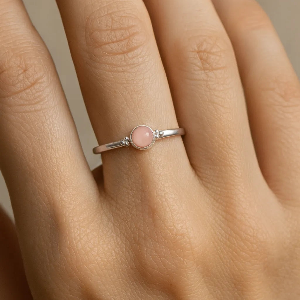 Nahaufnahme einer Frauenhand mit einem filigranen Silberring am Ringfinger. Der Ring ist mit einem kleinen, etwa 4 mm großen, runden Rosenquarz besetzt, der in einer glatten Zargenfassung sitzt. Der zart rosafarbene Stein wirkt leicht transparent und harmoniert mit dem silbernen Glanz des Rings. Die Haut ist warm getönt und das weiche Licht betont sowohl die feine Oberfläche des Rings als auch die natürliche Struktur der Haut.
