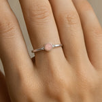 Nahaufnahme einer Frauenhand mit einem filigranen Silberring am Ringfinger. Der Ring ist mit einem kleinen, etwa 4 mm großen, runden Rosenquarz besetzt, der in einer glatten Zargenfassung sitzt. Der zart rosafarbene Stein wirkt leicht transparent und harmoniert mit dem silbernen Glanz des Rings. Die Haut ist warm getönt und das weiche Licht betont sowohl die feine Oberfläche des Rings als auch die natürliche Struktur der Haut.
