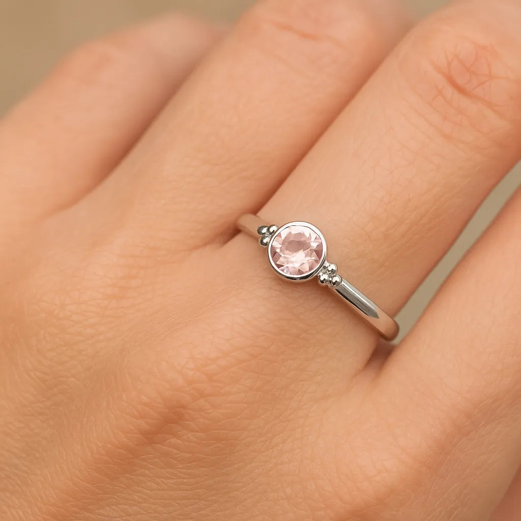 Eine Nahaufnahme einer Frauenhand zeigt einen filigranen Silberring mit einem zart roséfarbenen, facettierten Rosenquarz. Der runde Edelstein ist in eine glatte Fassung eingefasst und von kleinen silbernen Kugeldetails flankiert. Natürliches Licht lässt den Stein dezent funkeln und hebt den eleganten Glanz der polierten Ringschiene vor einem neutralen, unscharfen Hintergrund hervor.
