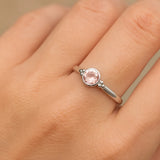 Eine Nahaufnahme einer Frauenhand zeigt einen filigranen Silberring mit einem zart roséfarbenen, facettierten Rosenquarz. Der runde Edelstein ist in eine glatte Fassung eingefasst und von kleinen silbernen Kugeldetails flankiert. Natürliches Licht lässt den Stein dezent funkeln und hebt den eleganten Glanz der polierten Ringschiene vor einem neutralen, unscharfen Hintergrund hervor.
