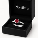 Ein silberner Ring mit einem runden, roten 4-mm-Stein liegt in einer geöffneten schwarzen Schmuckschatulle mit samtiger Einlage. Auf der Innenseite des Deckels steht das Logo „Novellara“ in heller Schrift. Das Bild ist in Studioqualität vor hellem Hintergrund aufgenommen.
