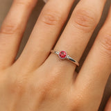 Nahaufnahme einer Frauenhand mit einem zarten Silberring am Ringfinger. Der Ring ist mit einem etwa 4 mm großen, facettierten Rubin besetzt, der in einer runden Zargenfassung sitzt. Kleine Silberkügelchen flankieren den Stein auf beiden Seiten. Das Licht hebt den Glanz des Silbers und das tiefe Rot des Rubins hervor, während die natürliche Hautstruktur der Hand fein sichtbar bleibt.
