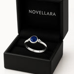 Ein silberner Ring mit einem runden, blauen 4-mm-Stein liegt in einer geöffneten schwarzen Schmuckschatulle mit samtigem Inlay. Auf der Innenseite des Deckels steht das Logo „NOVELLARA“ in heller Schrift. Das Bild ist in Studioqualität vor hellem Hintergrund aufgenommen.