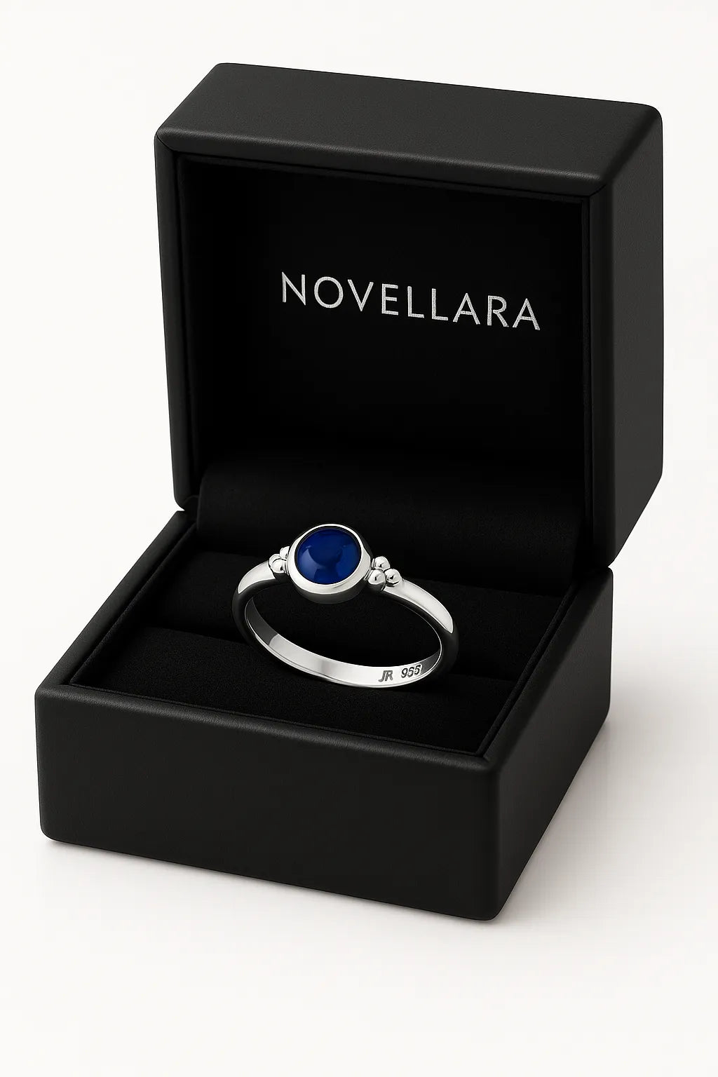 Ein silberner Ring mit einem runden, blauen 4-mm-Stein liegt in einer geöffneten schwarzen Schmuckschatulle mit samtigem Inlay. Auf der Innenseite des Deckels steht das Logo „NOVELLARA“ in heller Schrift. Das Bild ist in Studioqualität vor hellem Hintergrund aufgenommen.