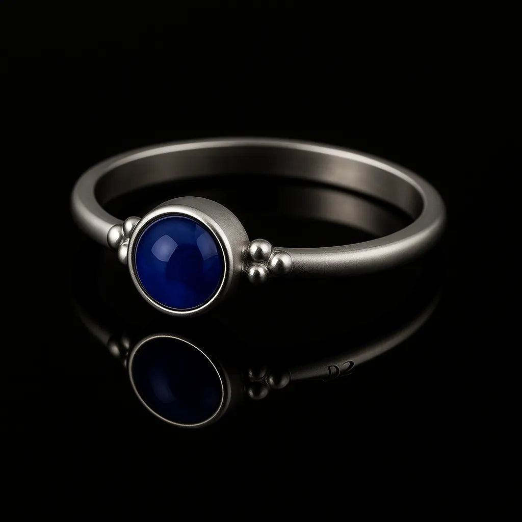 Eleganter Silberring mit rundem, glänzend dunkelblauem Stein in Zargenfassung und dekorativen Silberkügelchen an den Seiten, liegend auf schwarzem, hochglänzendem Untergrund mit deutlicher Spiegelung.