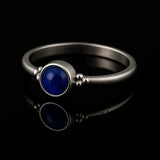 Eleganter Silberring mit rundem, glänzend dunkelblauem Stein in Zargenfassung und dekorativen Silberkügelchen an den Seiten, liegend auf schwarzem, hochglänzendem Untergrund mit deutlicher Spiegelung.