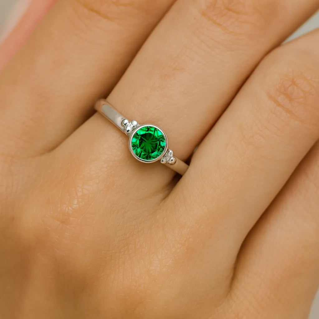 Nahaufnahme einer Frauenhand mit einem eleganten Silberring am Finger. Der Ring zeigt einen runden, facettierten Smaragd von etwa 4 mm Durchmesser, eingefasst in einer schlichten Zargenfassung mit kleinen Kugeldetails an den Seiten. Der grüne Edelstein funkelt lebhaft im Licht, während der Fokus klar auf dem Ring liegt und der Hintergrund weich verschwimmt.
