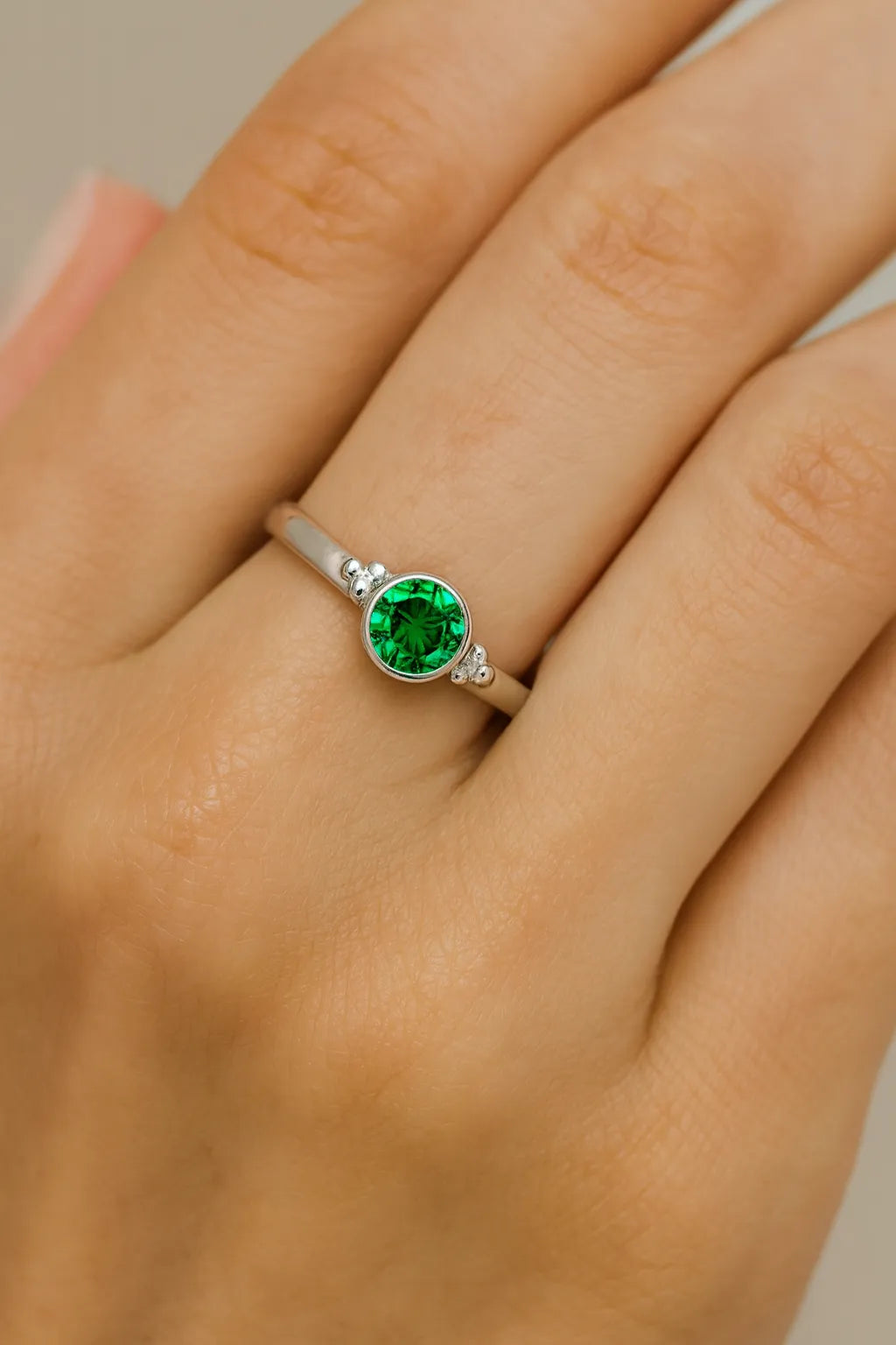 Nahaufnahme einer Frauenhand mit einem eleganten Silberring am Finger. Der Ring zeigt einen runden, facettierten Smaragd von etwa 4 mm Durchmesser, eingefasst in einer schlichten Zargenfassung mit kleinen Kugeldetails an den Seiten. Der grüne Edelstein funkelt lebhaft im Licht, während der Fokus klar auf dem Ring liegt und der Hintergrund weich verschwimmt.
