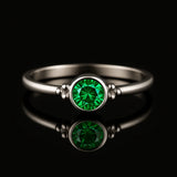 Damenring aus Sterling Silber mit rundem facettierten Smaragd in Zargenfassung – frontale Katalogansicht auf schwarzem spiegelnden Hintergrund, eleganter Silberring mit grünem Edelstein und feinen Silberkügelchen als edles Schmuckstück für Frauen.
