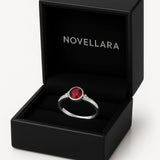 Ein silberner Ring mit einem runden, roten 4-mm-Stein liegt in einer offenen, schwarzen Schmuckschatulle mit Samteinlage. Auf der Innenseite des Deckels ist das Logo „NOVELLARA“ in heller Schrift zu sehen. Das Bild ist in hoher Studioqualität vor hellem Hintergrund aufgenommen.