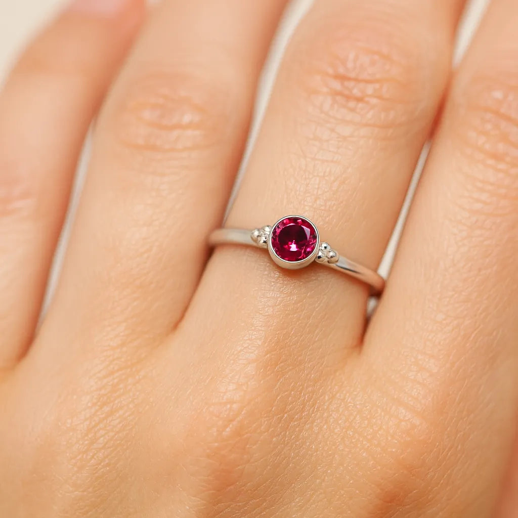 Nahaufnahme einer Frauenhand mit einem filigranen Silberring am Ringfinger. Der Ring besitzt einen kleinen, etwa 4 mm großen, facettierten roten Turmalin in einer runden Zargenfassung. Neben dem Stein befinden sich zwei kleine Silberkügelchen auf jeder Seite. Das weiche Licht hebt die glänzende Oberfläche des Silbers, die klare Facettierung des Edelsteins und die natürliche Struktur der Haut hervor.
