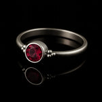 Schlichter Silberring mit rundem, facettierten, tief rubinroten Stein in Zargenfassung und dekorativen Silberkügelchen an den Seiten, liegend auf schwarzem, stark reflektierendem Untergrund mit deutlicher Spiegelung des Rings.
