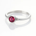 Feiner Silberring mit rundem, facettierten, intensiv pink- bis rubinroten Stein in Zargenfassung und kleinen Silberkügelchen an den Seiten, liegend auf hellem, spiegelndem Untergrund mit weicher Reflexion.