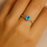 Eine Nahaufnahme einer Frauenhand zeigt einen eleganten Silberring mit einem facettierten, dreieckigen Blautopas auf dem Ringfinger. Der hellblau funkelnde Edelstein ist in einer glatten, modernen Fassung eingefasst, und das Licht betont seine klaren Facetten sowie das glänzende Silberband. Der Fokus des Bildes liegt auf dem Ring.
