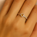 Nahaufnahme einer Frauenhand mit einem eleganten Silberring am Ringfinger. Der Ring besitzt einen dreieckigen, facettierten Citrinstein in warmem Goldgelb, der in einer glatten Zargenfassung sitzt. Das Licht spiegelt sich dezent auf der glänzenden Ringschiene, wodurch der klare, moderne Stil des Schmuckstücks betont wird. Der Fokus liegt deutlich auf dem Ring, während die Hand einen natürlichen Hautton aufweist und weich beleuchtet ist.
