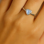 Eine Nahaufnahme einer Frauenhand zeigt einen eleganten Silberring am Ringfinger. Der Ring besitzt eine schlichte, glatte Ringschiene und ist mit einem dreieckig facettierten, hellblauen Blautopas von etwa 5 mm Länge besetzt. Der Fokus liegt klar auf dem Ring und seinem funkelnden Edelstein, während die Haut im Hintergrund weich und natürlich wirkt.
