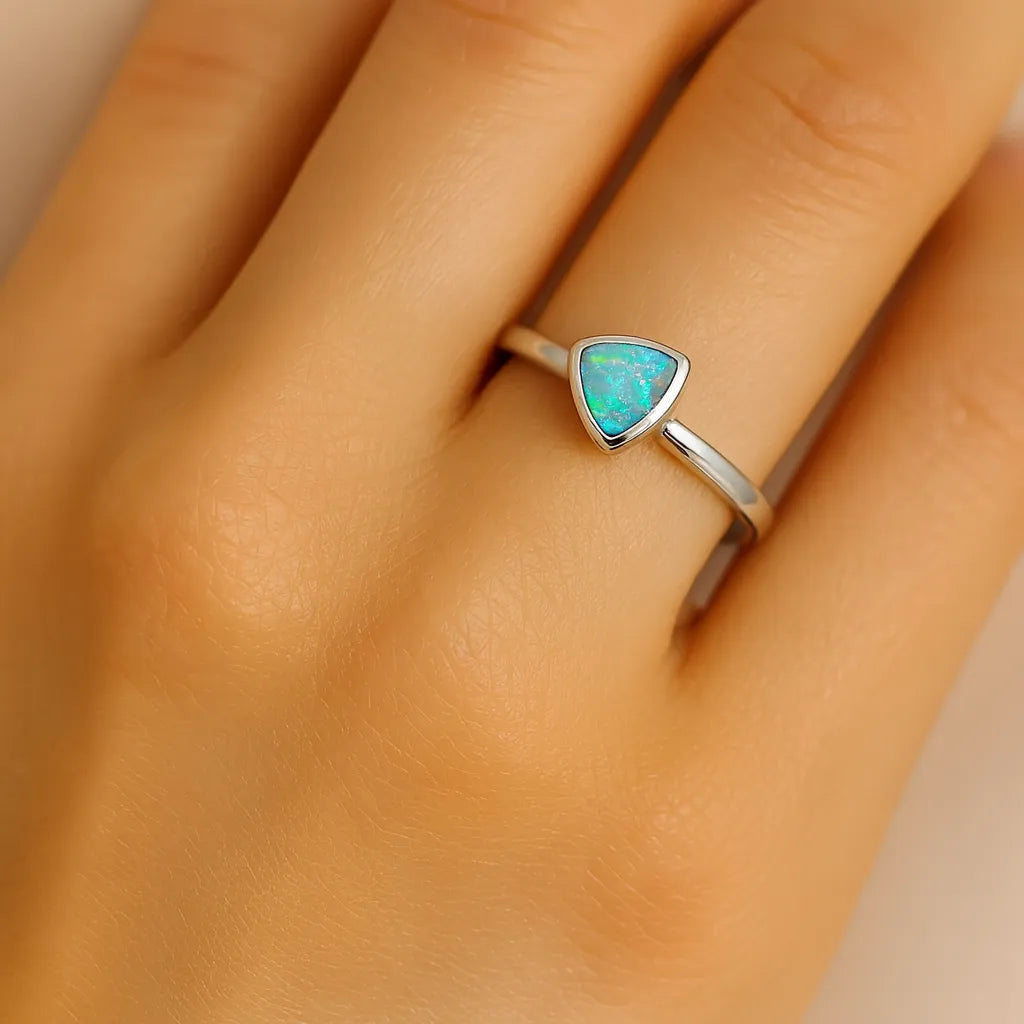 Nahaufnahme einer Frauenhand mit einem eleganten Silberring am Ringfinger. Der Ring besitzt eine zarte, glatte Ringschiene und einen kleinen, dreieckigen Opal in einer schlichten Fassung. Der Edelstein schimmert in dezenten Blau-, Grün- und Orangetönen, während der Fokus des Bildes klar auf dem Ring liegt.
