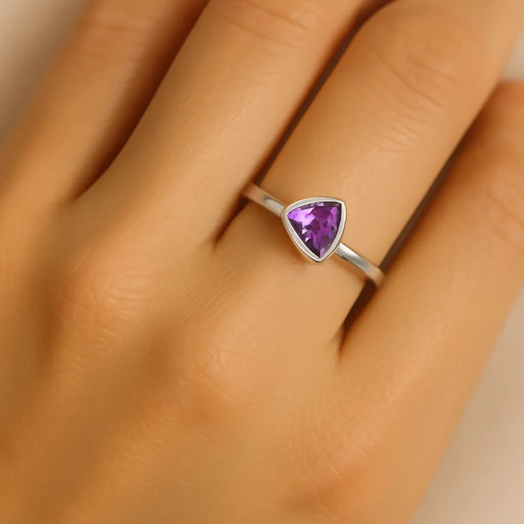 Eine Nahaufnahme zeigt die linke Hand einer Frau mit einem eleganten Silberring am Ringfinger. Der Ring besitzt eine feine, dreieckige Fassung mit einem facettierten, violetten Amethyst als Stein. Der Fokus liegt auf dem Ring, während die Hand weich beleuchtet ist und einen natürlichen Hautton zeigt.
