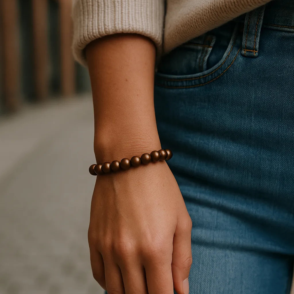 Eine Person trägt ein bronzefarbenes Perlenarmband am Handgelenk. Der Fokus liegt auf dem Armband, im Hintergrund ist eine unscharfe urbane Straßenszene erkennbar.