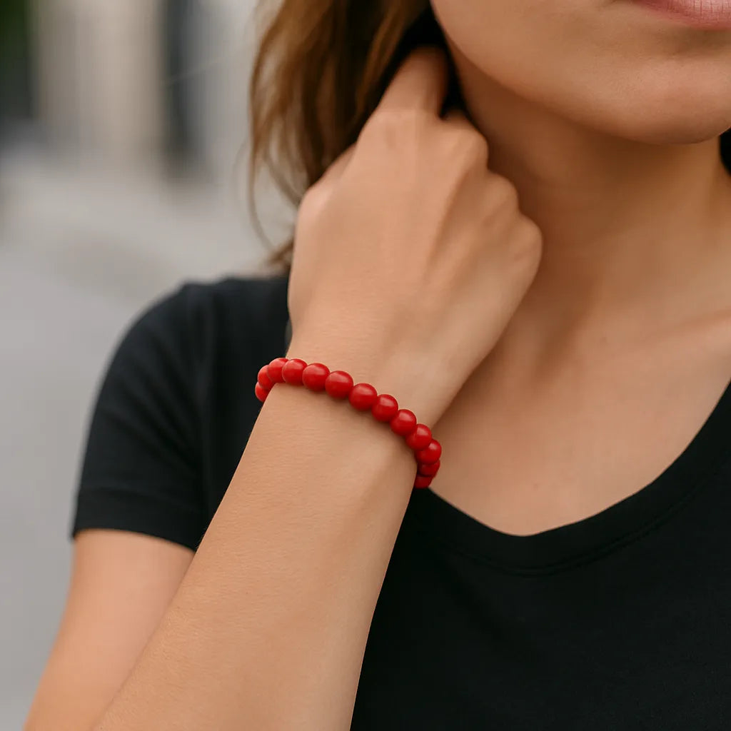 Eine Frau trägt ein rotes Perlenarmband am Handgelenk. Der Fokus liegt auf dem Armband, während sie ihre Hand locker zum Hals führt. Der Hintergrund ist unscharf urban gehalten.